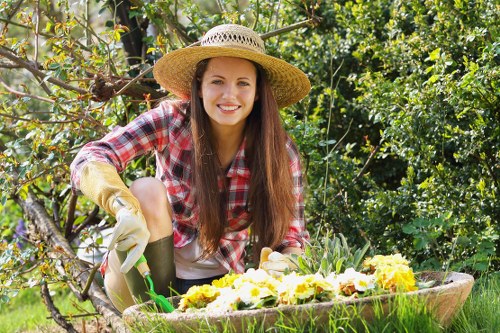 A young woman with long brown hair, wearing a straw sunhat, a plaid long-sleeve shirt, beige gardening gloves, and light-colored trousers, is kneeling in a backyard garden during daytime. She is planting or tending to a variety of blooming flowers arranged in a large, shallow wooden or ceramic container. The garden behind her features a lush hedge and flowering shrubs with green leaves and some yellow blossoms, indicating a well-maintained outdoor space typical of a residential garden in Hackney. The scene shows bright natural sunlight and a clear sky, highlighting the vibrant colors of the flowers, foliage, and the woman's clothing. The overall setting depicts a neat, designed garden area with a lawn in the foreground, and the outdoor environment suggests ongoing gardening or landscaping work, consistent with professional gardening services such as hedge trimming and planting care provided by companies like Hedge Trimming Hackney.