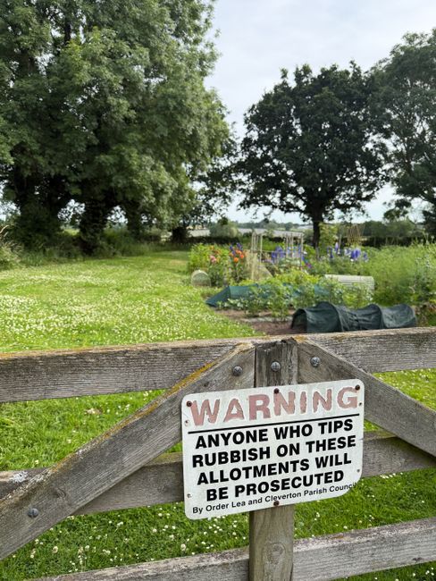 A weathered wooden gate with a warning sign attached, reading 'WARNING: Anyone who tips rubbish on these allotments will be prosecuted by the Cleventon Parish Council, ordered by Lea and Cleventon Par